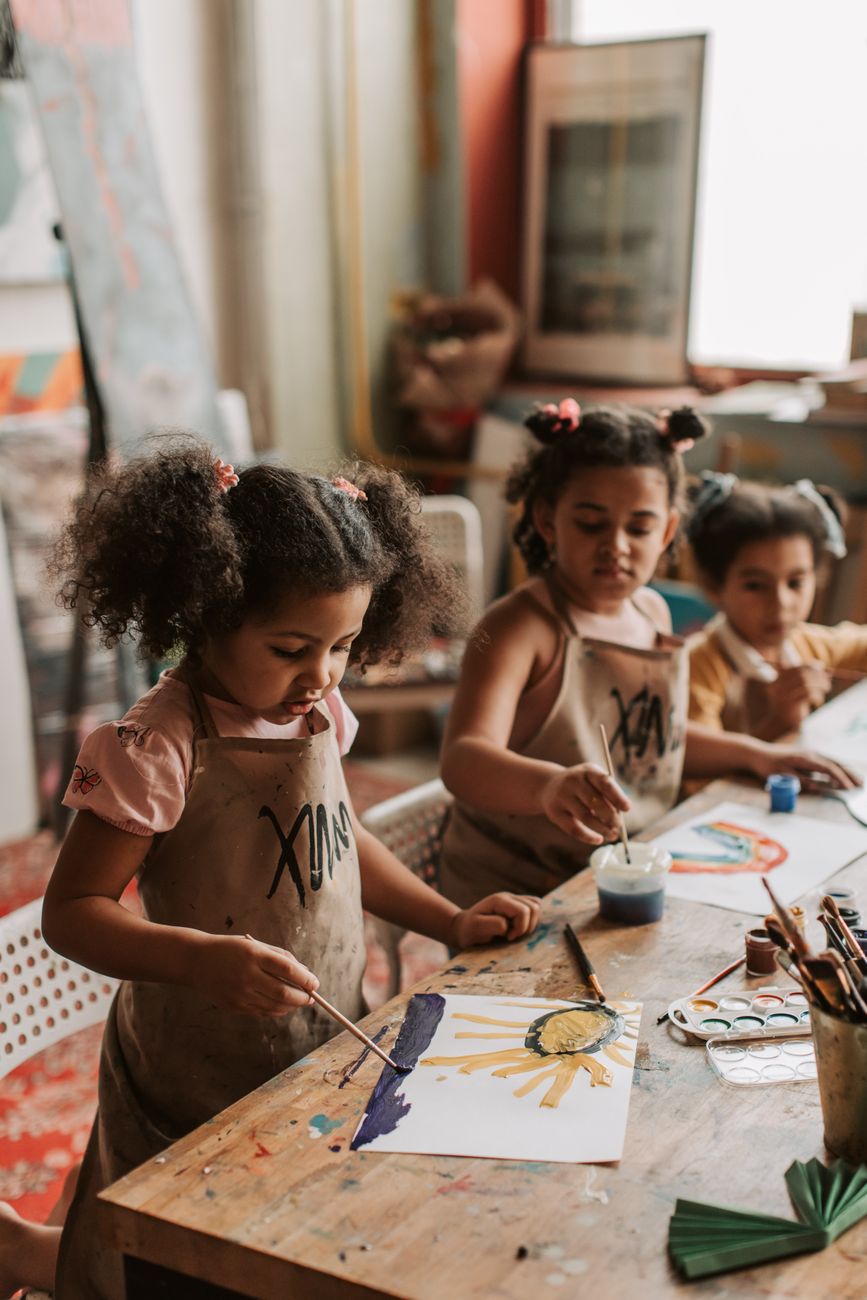Plate 03. Three children in aprons paint at a wooden atelier table, each on a different sheet.