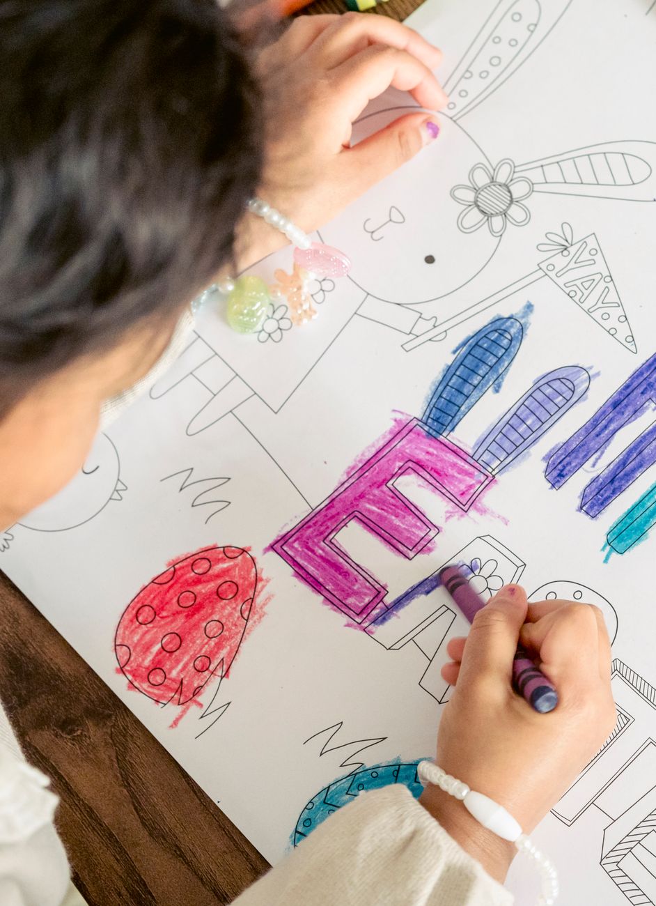 Plate 06. A child colours in a large lettered poster with wax crayons, a teacher's hand guiding the paper.