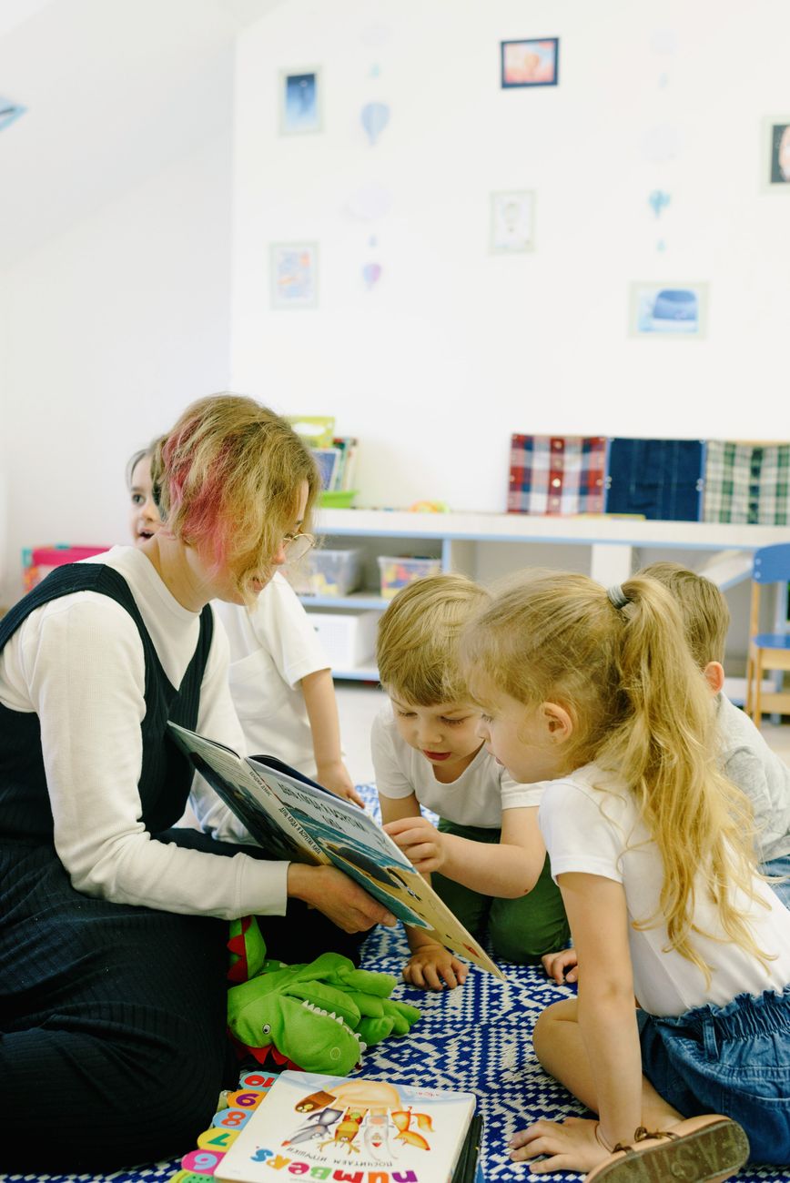 An educator reads a picture book to a group of children seated on a rug beneath framed drawings.