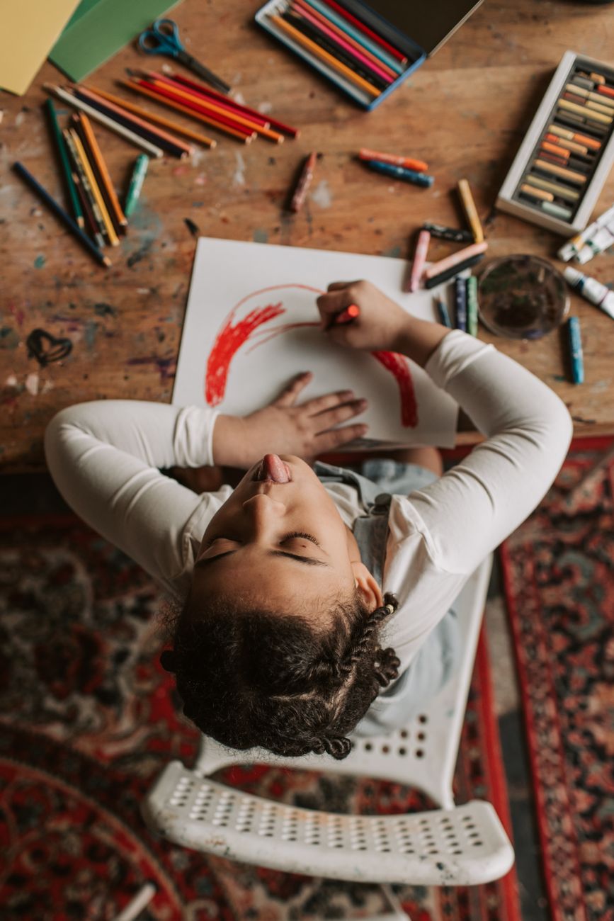 Plate 11. Seen from above, a girl draws a red arc on white paper among crayons and pastels.