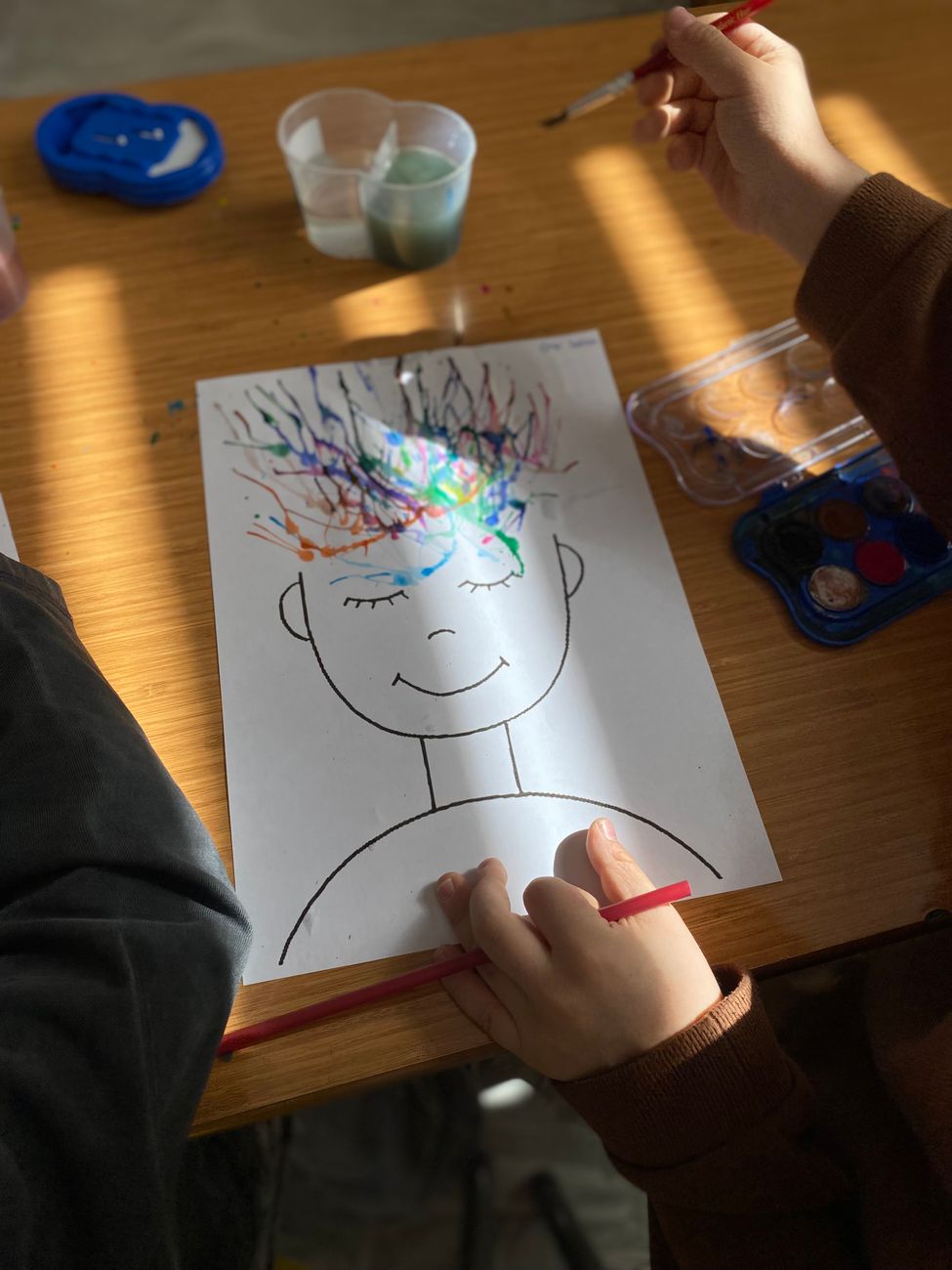 Plate 01. A child draws a face and paints radiating colour above it while another hand holds a brush, mid-project on a sunlit table.