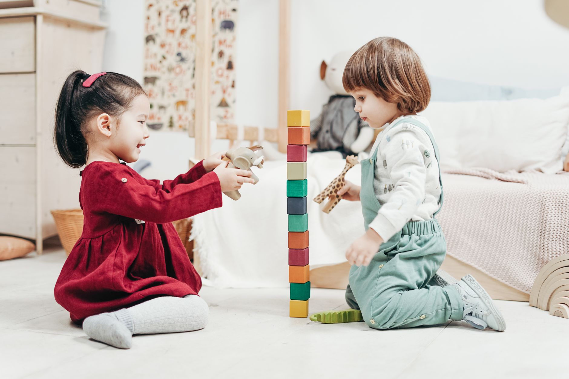 Two young children build a tall tower of coloured blocks on a bright playroom floor.