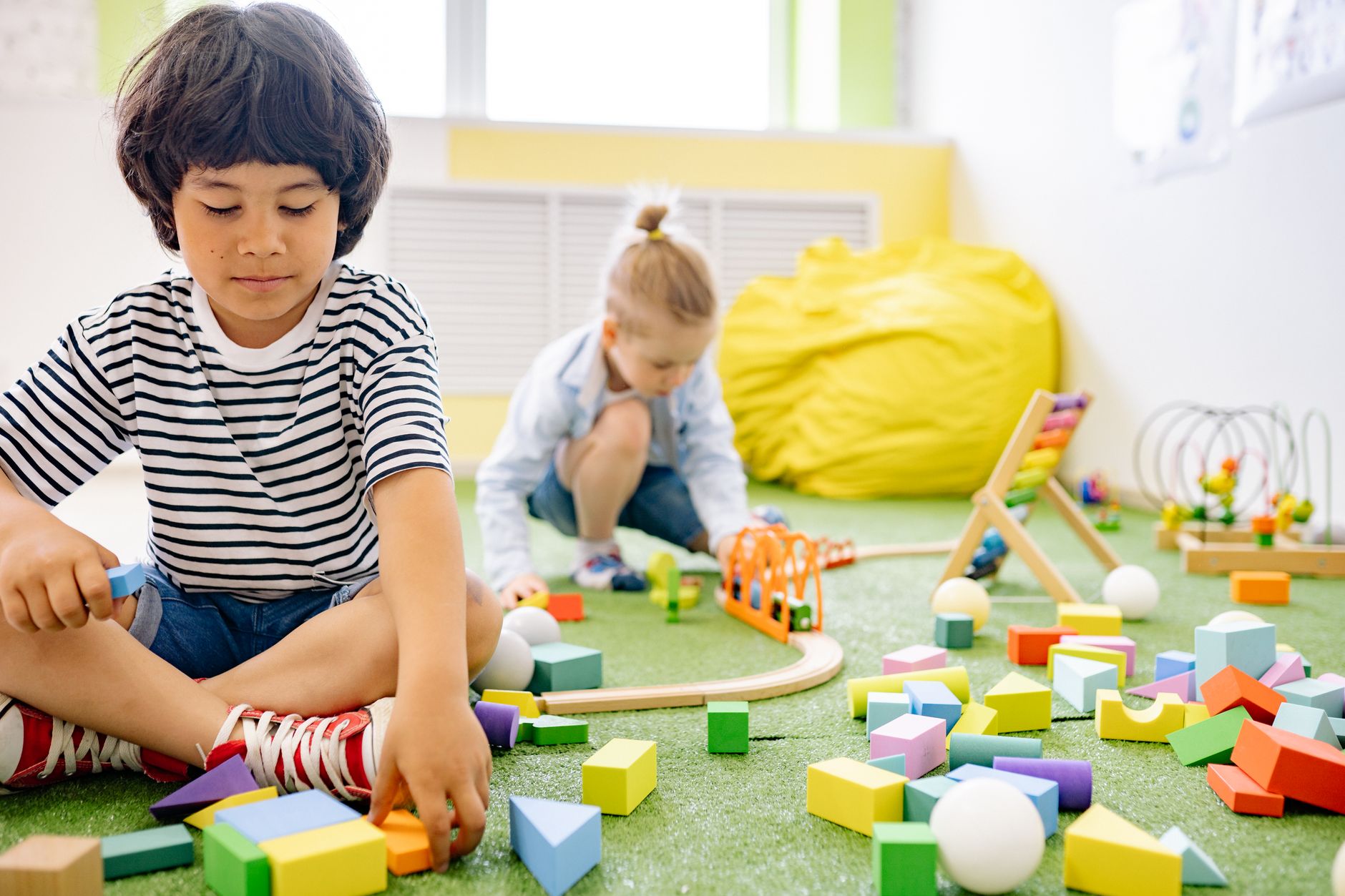A boy in a striped shirt builds with shapes and a wooden train track while a friend plays beyond.