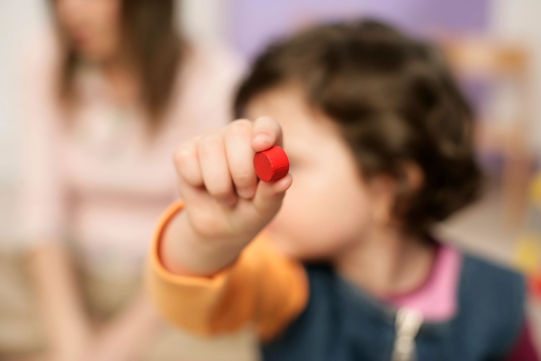 A toddler holds up a small red block, other children soft-focus behind.