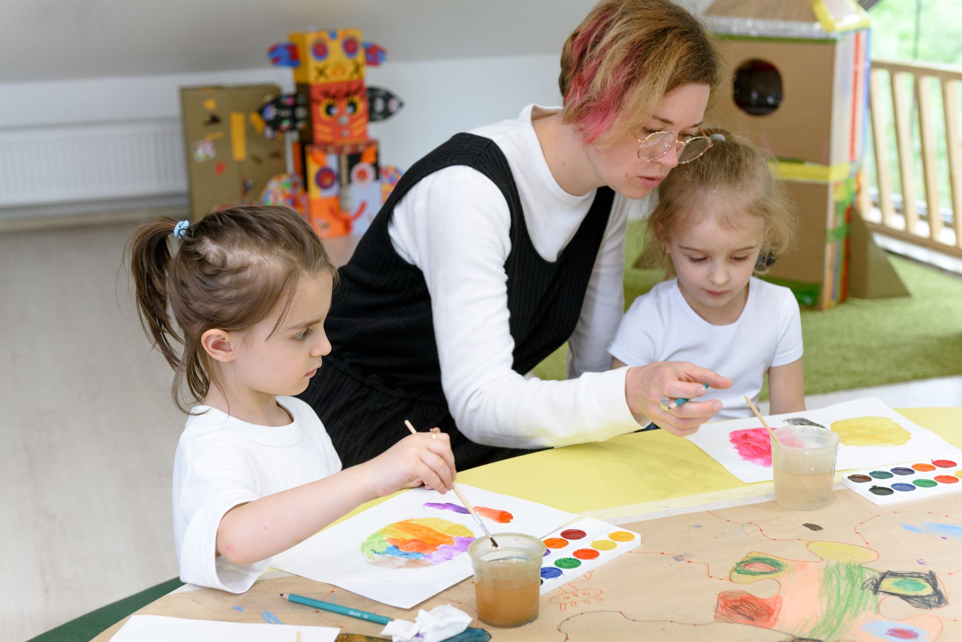 An educator and a girl paint together with watercolours beside a cardboard construction.